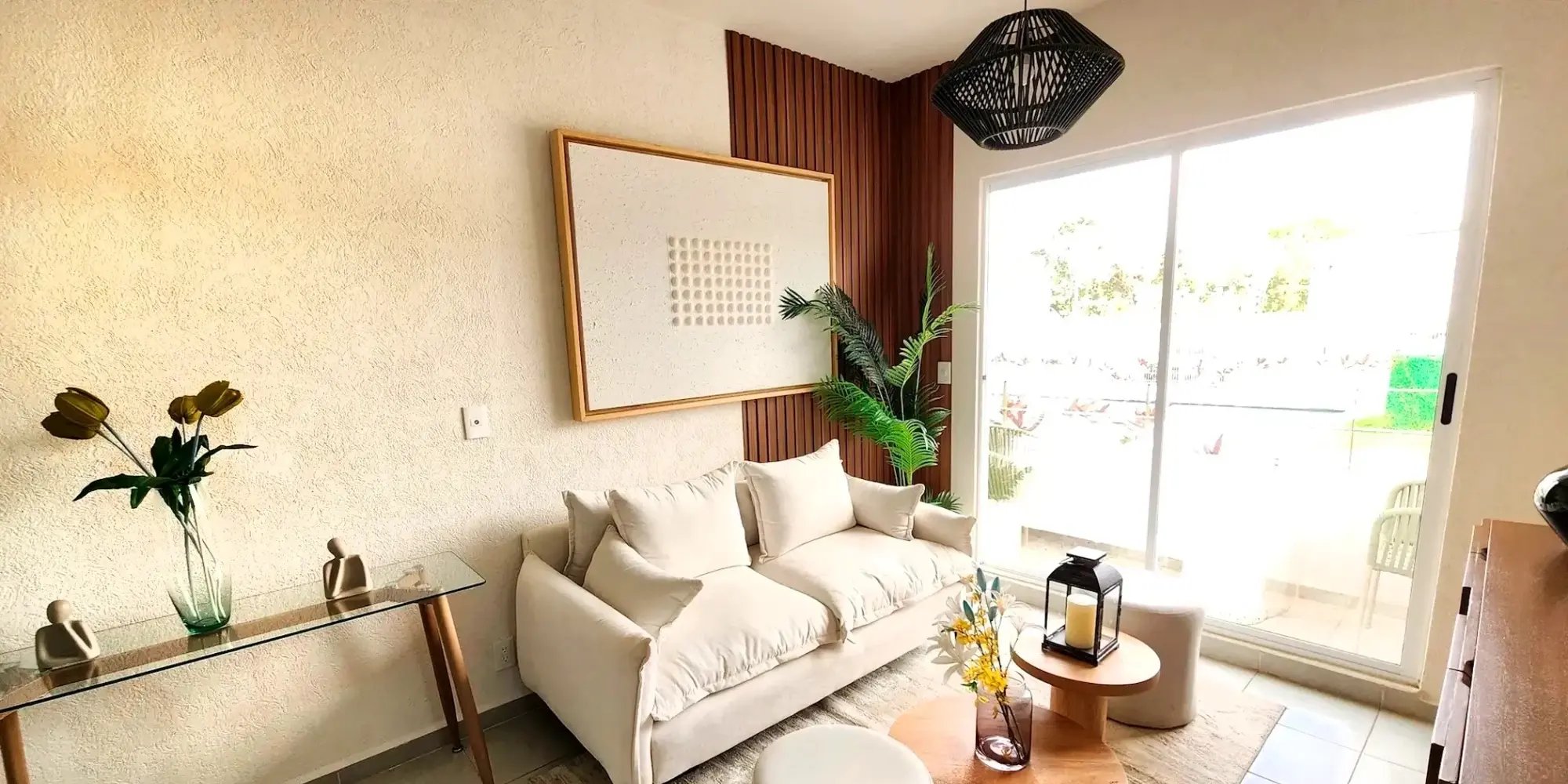 Sala moderna con decoración minimalista, sofá blanco, mesa de centro de madera y elementos decorativos como flores y figuras humanas. Espacio luminoso con vistas a la piscina.