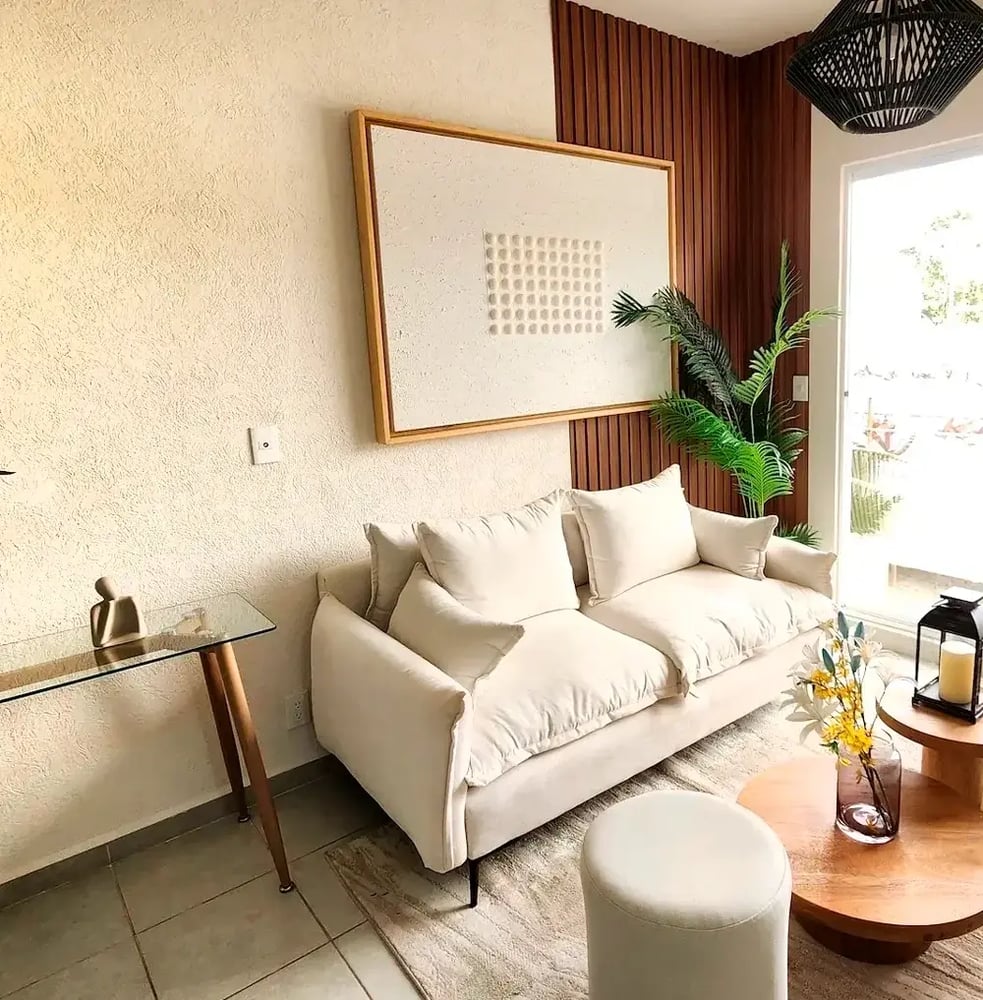 Sala moderna con decoración minimalista, sofá blanco, mesa de centro de madera y elementos decorativos como flores y figuras humanas. Espacio luminoso con vistas a la piscina.